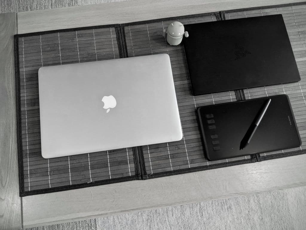 A closed macbook pro, a modern laptop, a graphics tablet and an Android toy are on a wooden coffee table.